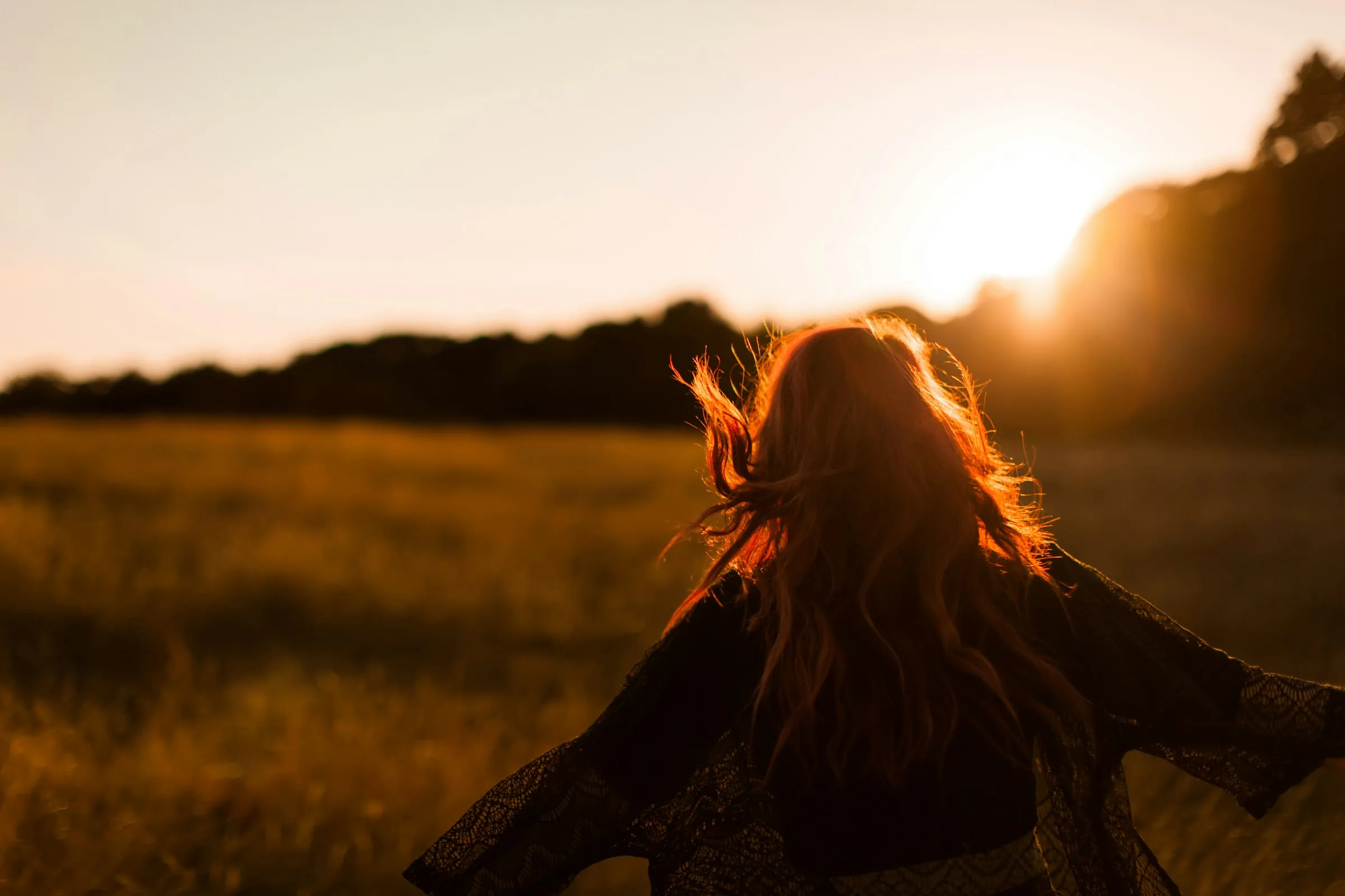 A woman with her back turned, arms open to a low golden sun over a field